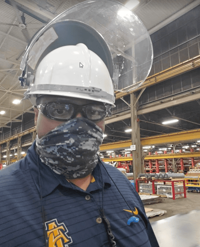 A man wearing a hardhat with face shield, safety glasses and a gaiter looks at the camera.